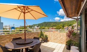 Eine Terrasse mit einem runden Tisch, Stühlen, einem beigen Sonnenschirm und Blumentöpfen.