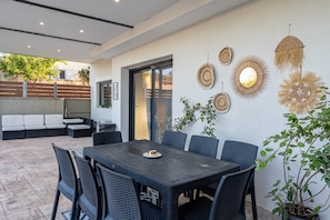 Un coin repas avec une table en bois foncé et des chaises en osier, un mur blanc orné d’une décoration murale tissée et une plante en pot.