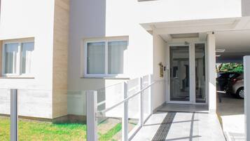 A building entrance with a glass door, a small outdoor area with a metal railing, and a parked car in the background.