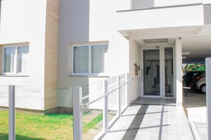 Una entrada de edificio con puerta de cristal, un pequeño espacio exterior con barandilla metálica y un coche estacionado al fondo.
