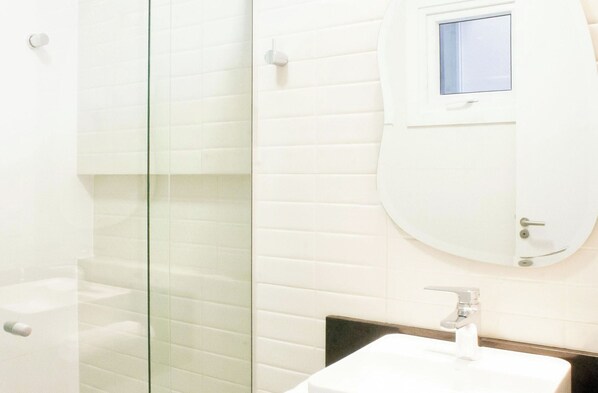 Un baño con cabina de ducha de vidrio, un lavamanos blanco y un espejo con un pequeño ventanal.