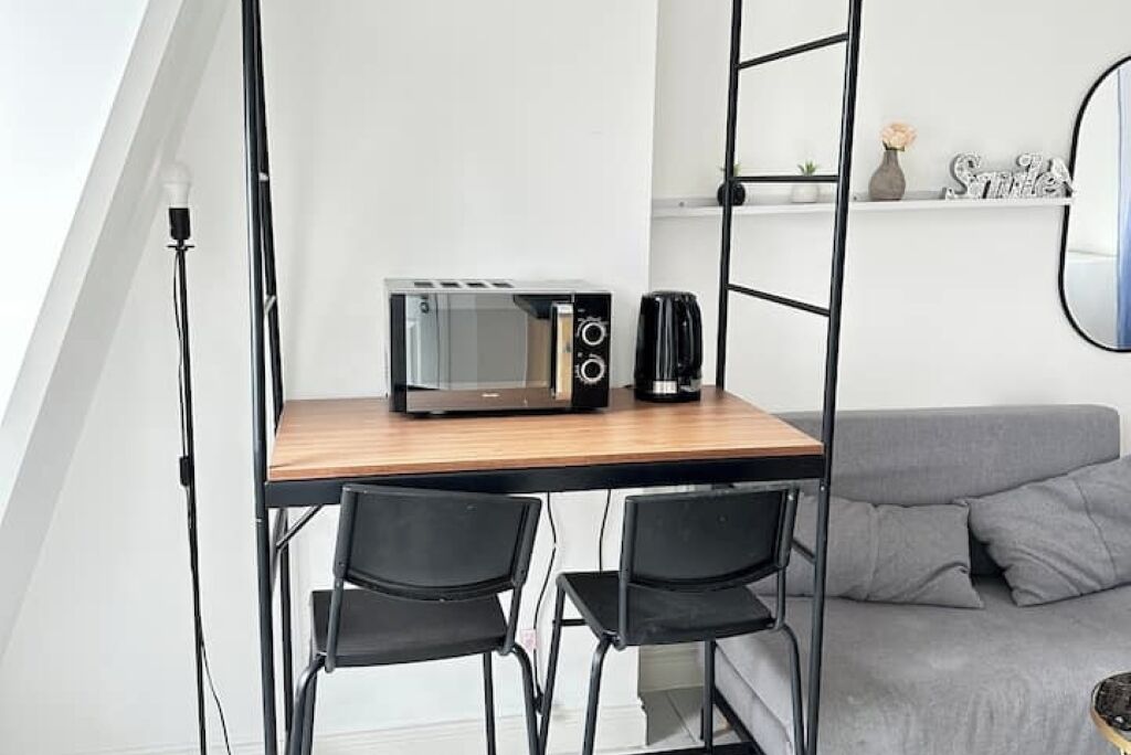 A compact kitchenette with a microwave, toaster, and coffee maker on a wooden table, two black chairs, and a grey sofa in a modern apartment.