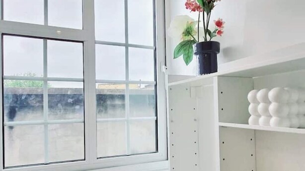Een kamer met een groot raam, een plant in een pot op een plank en witte planken met decoratieve objecten.