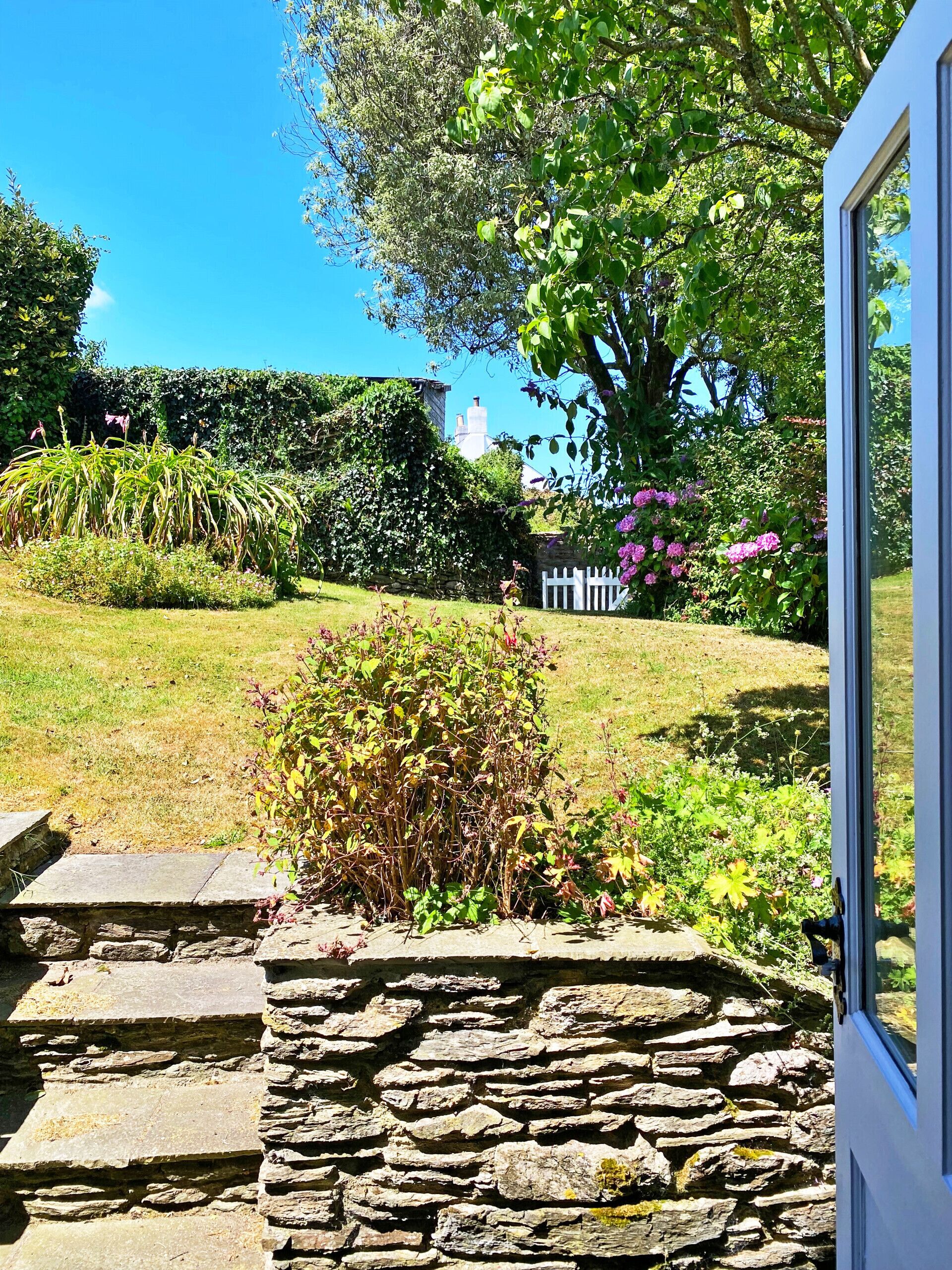 A stone pathway leading to a white fence, with a garden featuring various plants and a large tree in the background.