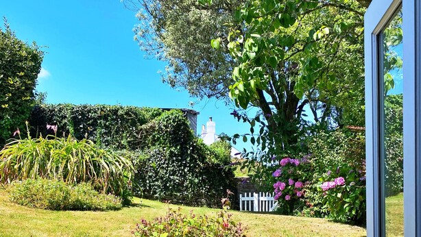 A stone pathway leading to a white fence, with a garden featuring various plants and a large tree in the background.