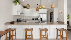A modern kitchen with a central island, white cabinetry, stainless steel appliances, and three wooden stools. - Beachfront Condo In Costa Rica (Tempate)