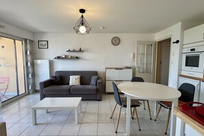 En moderne stue med sofa, sofabord, spisebord og stole. Der er et køkkenområde med skabe og et køleskab.