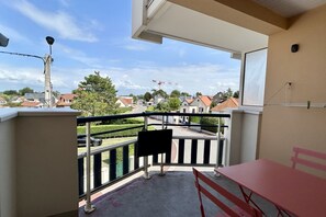 Un balcón con una mesa y un banco rojos, vista a la calle con casas y una grúa, y cielo despejado.