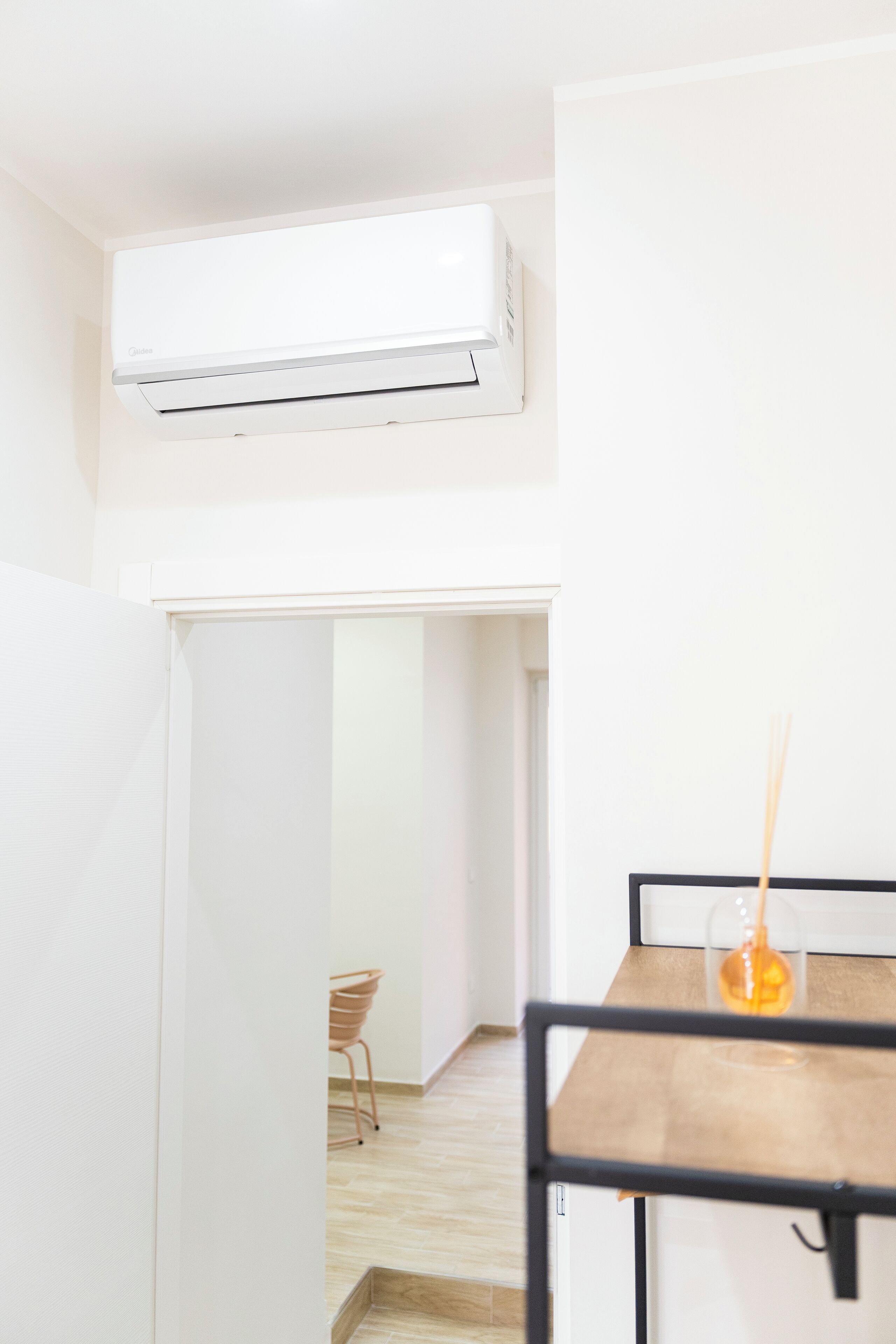 A modern, minimalistic room with a white air conditioning unit, a wooden table with a glass vase, and a chair in the background.
