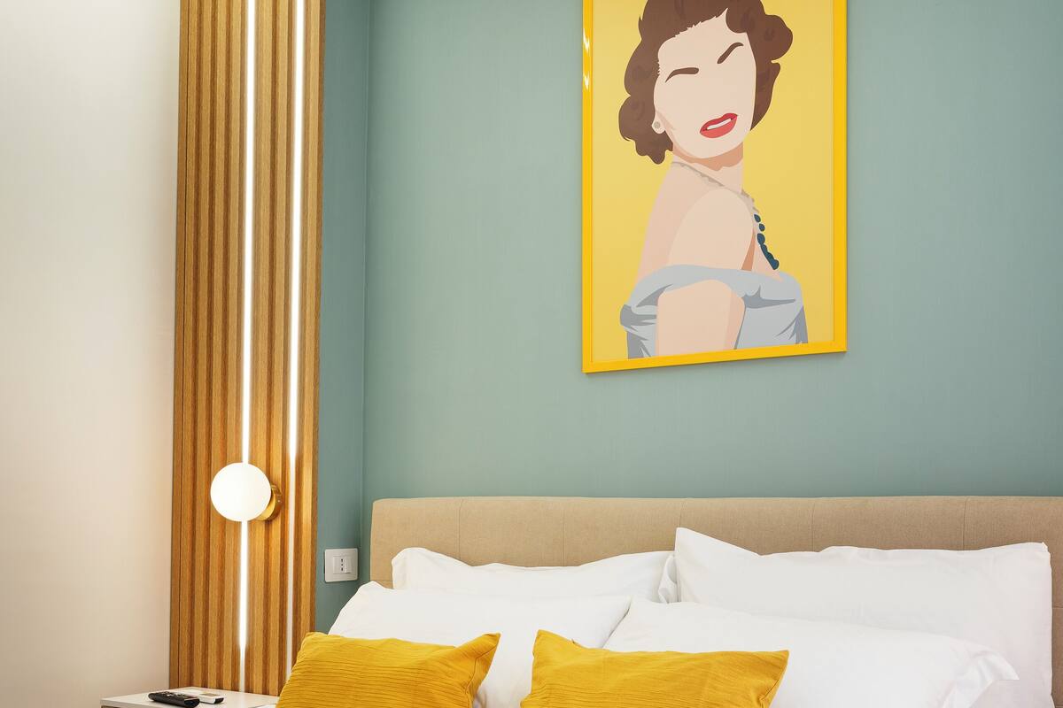 A neatly made bed with white and mustard pillows, a wooden panel on the wall, and a framed artwork above the bed.