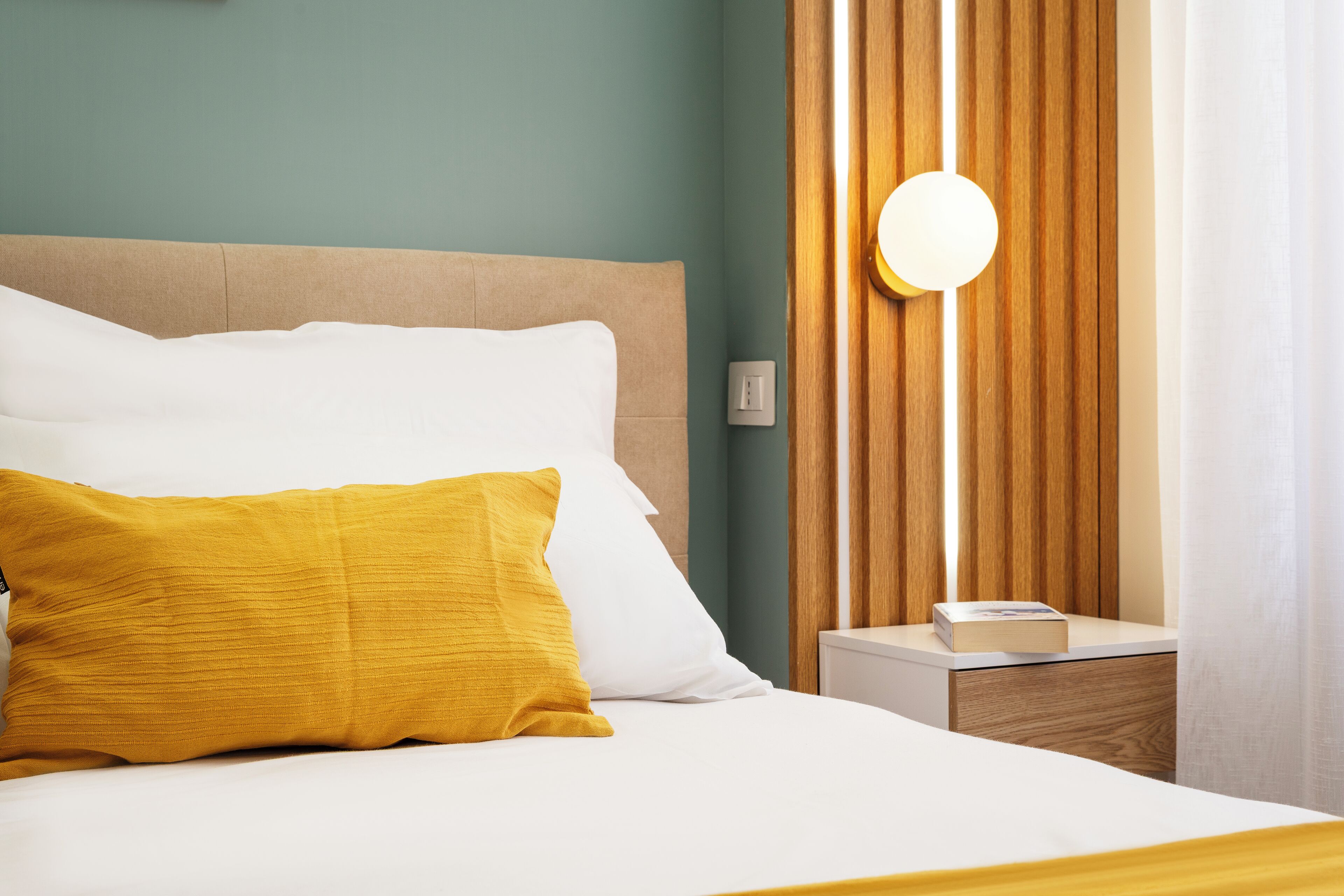 A neatly made bed with white and mustard pillows, a wooden headboard, a bedside table with a book, and a wall-mounted light.