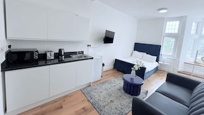 A compact living space with a kitchenette, a bed with a dark headboard, a small round table, and a sofa. - Studio Flat 2 in Golders Green (London)