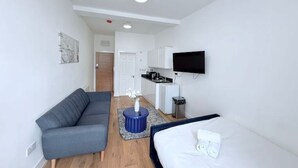 A modern, compact room with a grey sofa, a bed, a small round table with a vase, a TV mounted on the wall, and a kitchenette in the background. - Studio Flat 2 in Golders Green (London)