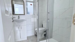 A bathroom with a white sink, mirror, and toilet, and a glass-enclosed shower area. - Studio Flat 2 in Golders Green (London)