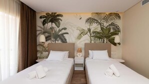 A hotel room with two single beds, a nightstand with a lamp, and a tropical-themed mural on the wall.