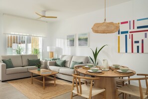 A modern living room with a sofa, coffee table, dining table, and chairs. There are plants, artwork on the wall, and a ceiling fan.