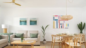 Una sala de estar moderna con sofá, mesa de centro y zona de comedor. Hay dos cuadros en la pared, un ventilador de techo y una planta de cactus.