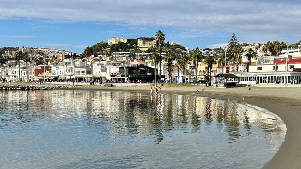 Un paisaje costero con playa, edificios y una colina con una estructura similar a un castillo.