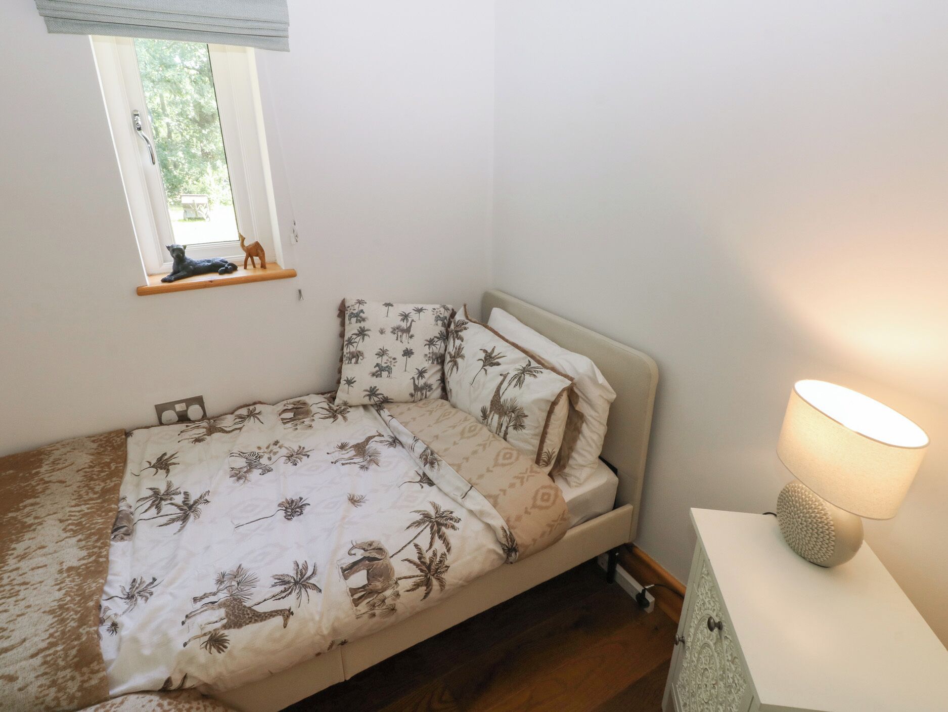 A small bedroom with a single bed, a nightstand, a lamp, a window with a view of trees, and a shelf with a cat figurine.