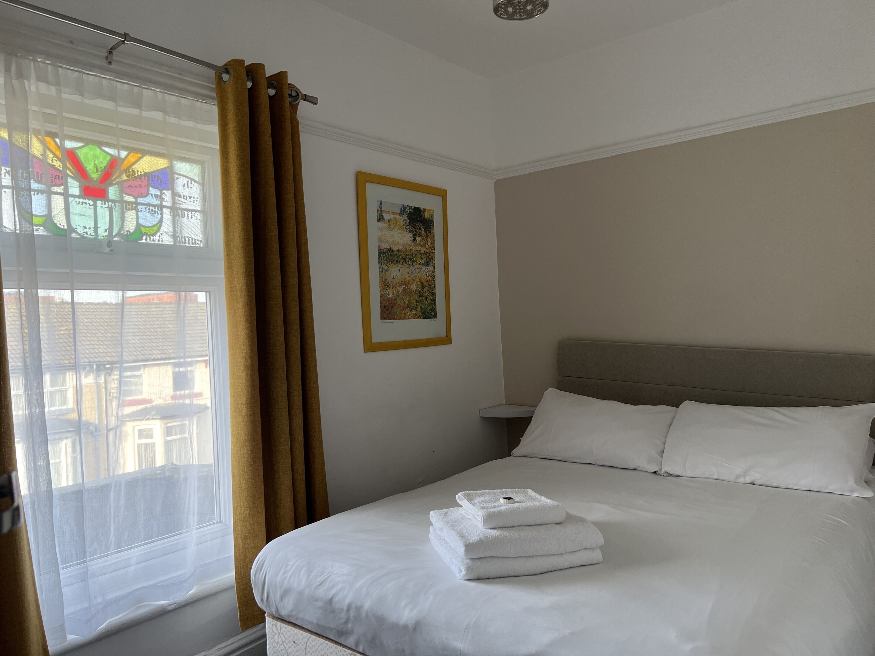 A hotel room with a bed, a window with curtains, a framed picture on the wall, and a view of buildings outside.