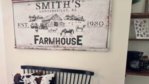 A rustic wall hanging with a farmhouse illustration, a black metal bench with cowhide cushions, and a woven basket. - Adorable 2-bedroom Farmhouse in beautiful Lake Guntersville (Guntersville)