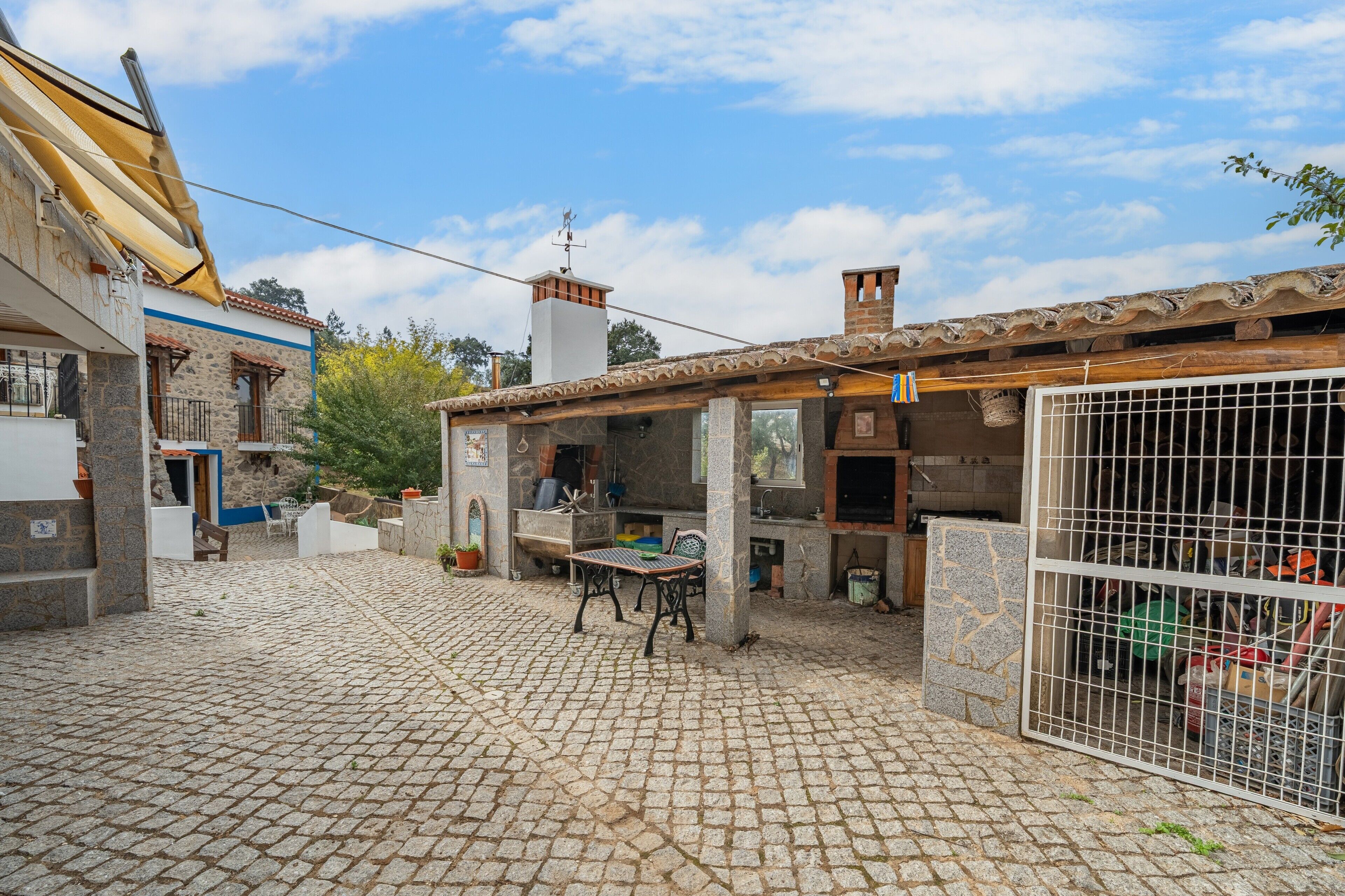 A cobblestone courtyard with a table and chairs, a fenced area with various items, and buildings with stone and tiled roofs.