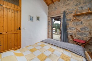 Un dormitorio con puerta de madera, cama con colcha a cuadros, pared de piedra y silla de madera con cojín rojo.