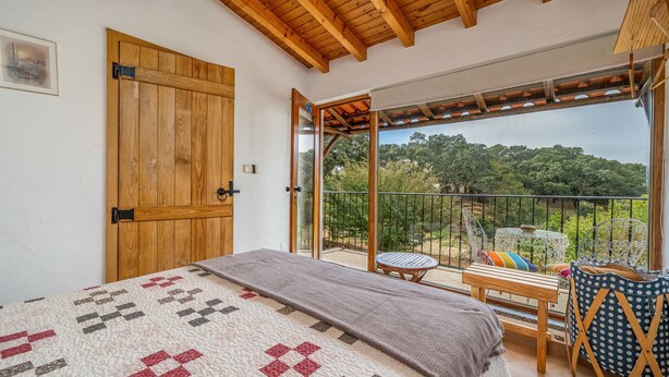 A bedroom with a large bed, a wooden door, a balcony with a table and chairs, and a view of greenery.