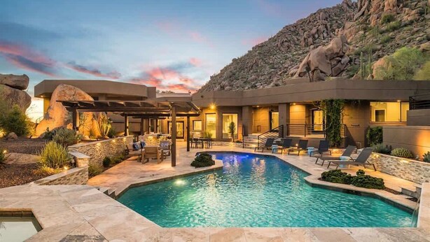 Zona de piscina con cubierta de piedra, sillas de descanso y un edificio con porche cubierto.