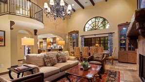 A spacious living area with a high ceiling, a chandelier, a large window with a view, a fireplace, and a dining area with chairs and a table.