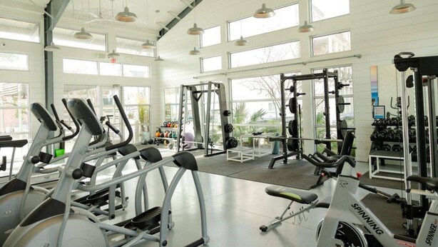 A spacious gym with various exercise equipment, large windows, and a high ceiling.