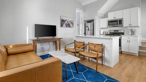 A modern living room with a sofa, a coffee table, and a TV. There is a kitchen with white cabinets, a microwave, and a refrigerator.