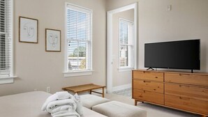 Une chambre à coucher comprenant un lit, une commode en bois, un téléviseur et deux fenêtres équipées de stores.