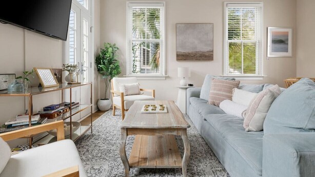 Un salon avec un canapé, une table basse et un téléviseur fixé au mur. On y trouve des fenêtres équipées de stores, une plante et un tableau accroché au mur.