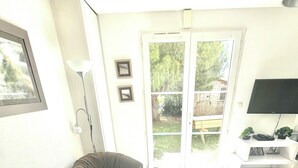 Una sala de estar con dos sillones de peluche, un televisor de pantalla plana sobre un pedestal blanco y una puerta que da a un jardín.