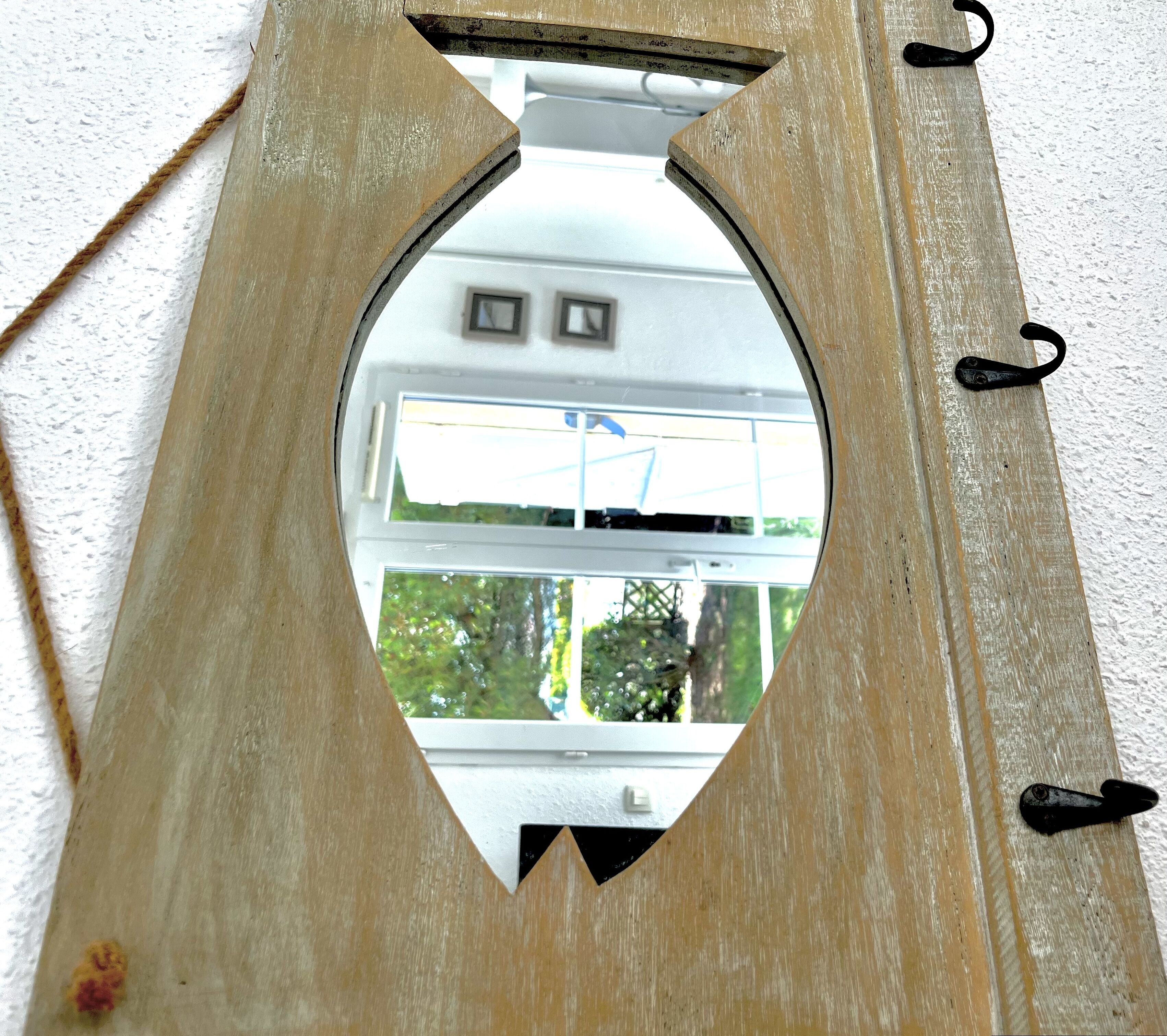 Ein spiegel im Fischdesign aus Holz mit Haken an der Unterseite, an einer weißen Wand befestigt.