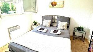 Un dormitorio con cama, ventana, radiador, una mesita con una planta y una obra de arte colgada en la pared.