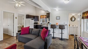 Ein modernes Wohnzimmer mit einem grauen Ecksofa, Deckenventilator und einer Küchenzeile mit weißen Schränken und einer Mikrowelle.