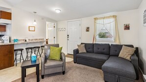 Ein modernes Wohnzimmer mit einem grauen Sofa, einem Ledersessel und einem kleinen Tisch. Es gibt eine Küchenzeile mit einem Barhocker und einer Arbeitsfläche.