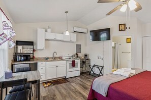 A compact hotel room with a kitchenette, a bed with a red blanket, a ceiling fan, and a TV mounted on the wall.