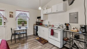 A compact kitchen with white cabinets, a stove, a microwave, and a coffee maker. There is a dining table with chairs and a window with curtains. - Merlot Cottage - GOTL Strip - Lover's Retreat (Geneva)