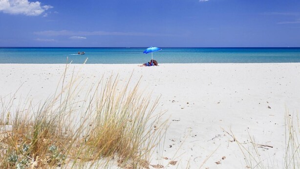 Une plage de sable blanc, un ciel bleu azur et une personne abritée sous un parasol bleu.