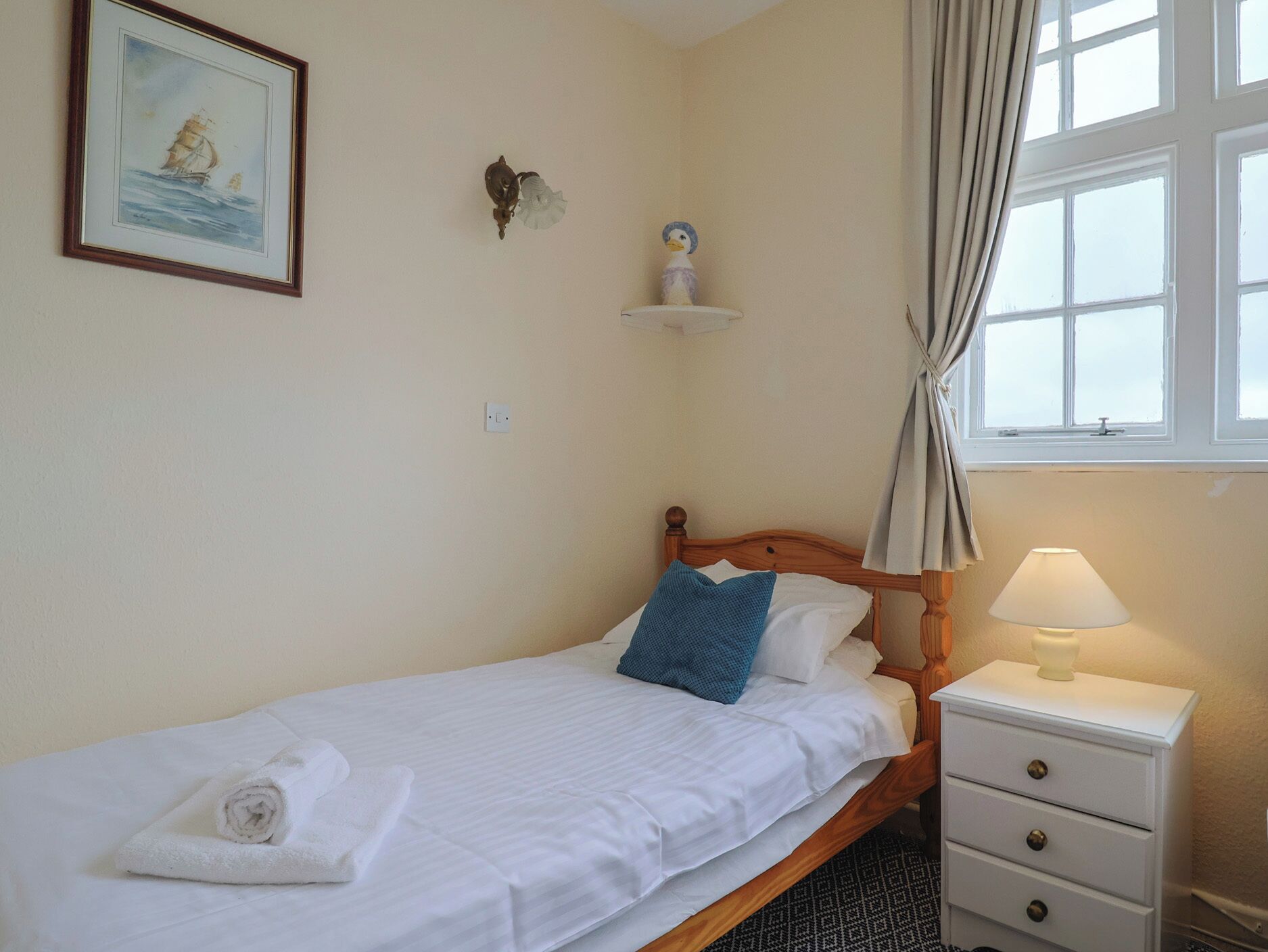 A neatly made bed with white linens, a blue pillow, and a rolled towel on the floor. There is a wooden nightstand with a lamp and a small shelf above it. A framed picture hangs on the wall, and a window with curtains is visible.