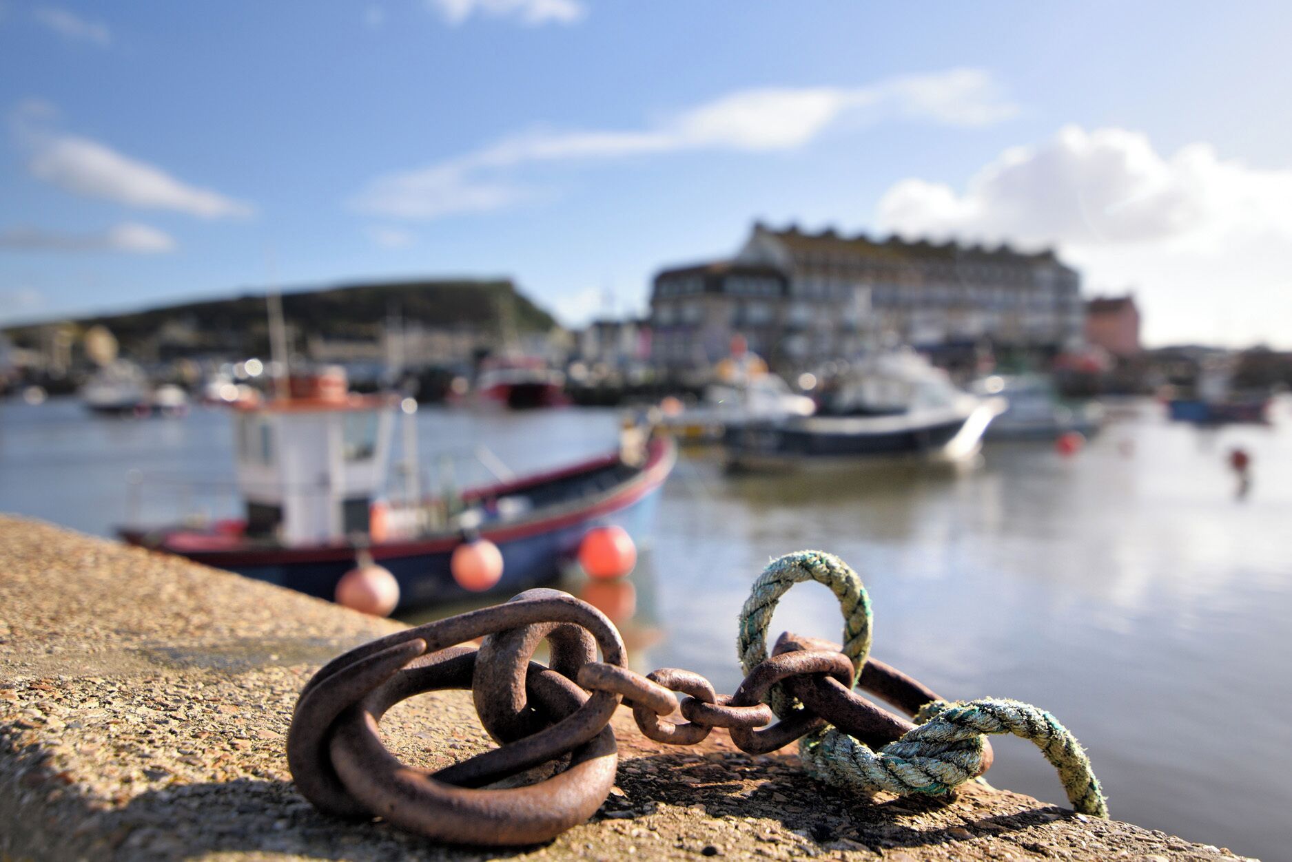Gros plan d’une chaîne rouillée et d’une corde attachées à une surface en béton, avec un port et des bateaux en arrière-plan.
