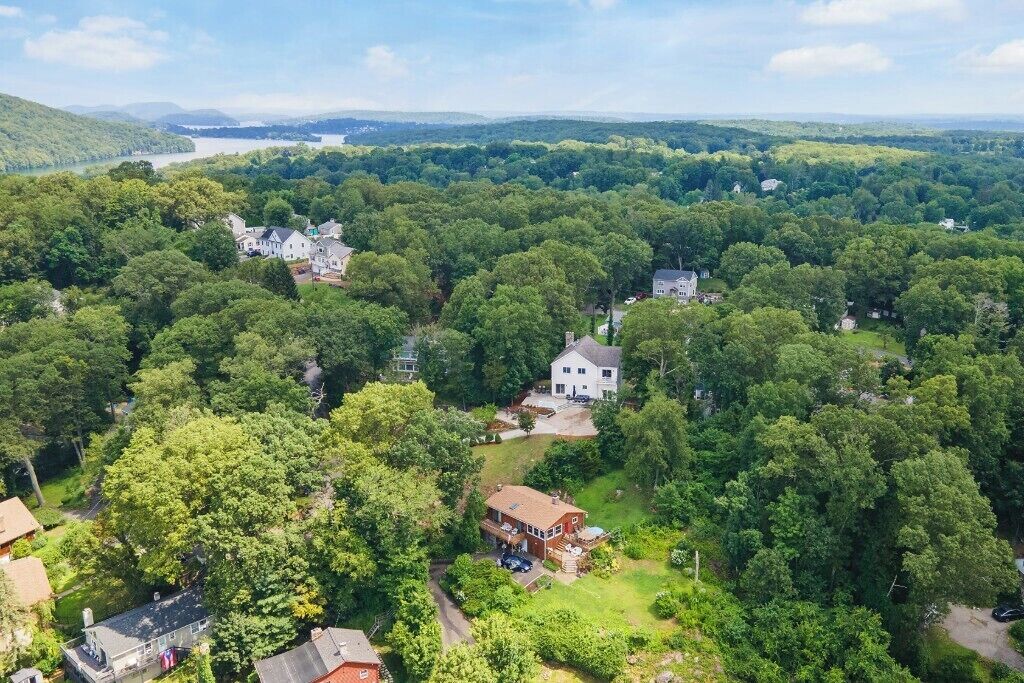 Luchtbeeld van een woongebied met meerdere huizen, omgeven door dicht bos, een meer op de achtergrond en een heldere lucht.