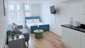 A modern, well-lit room with a bed, a sofa, a small table with a plant, a round side table, a TV mounted on the wall, and a kitchen area with cabinets and appliances.
