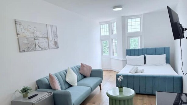 A modern room with a blue sofa, a bed with a blue headboard, a small green table, and a flat-screen TV mounted on the wall.