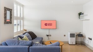 Moderni olohuone, jossa on harmaa sohva, litteänäyttöinen televisio, jossa näkyy teksti ”VALMIS VIERAITA VARTEN”, ja pieni pöytä, jonka päällä on kasvi.