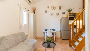 Um espaço de estar compacto com sofá, uma mesa de jantar com cadeiras, uma pequena cozinha com frigorífico e micro-ondas, e uma escadaria de madeira.