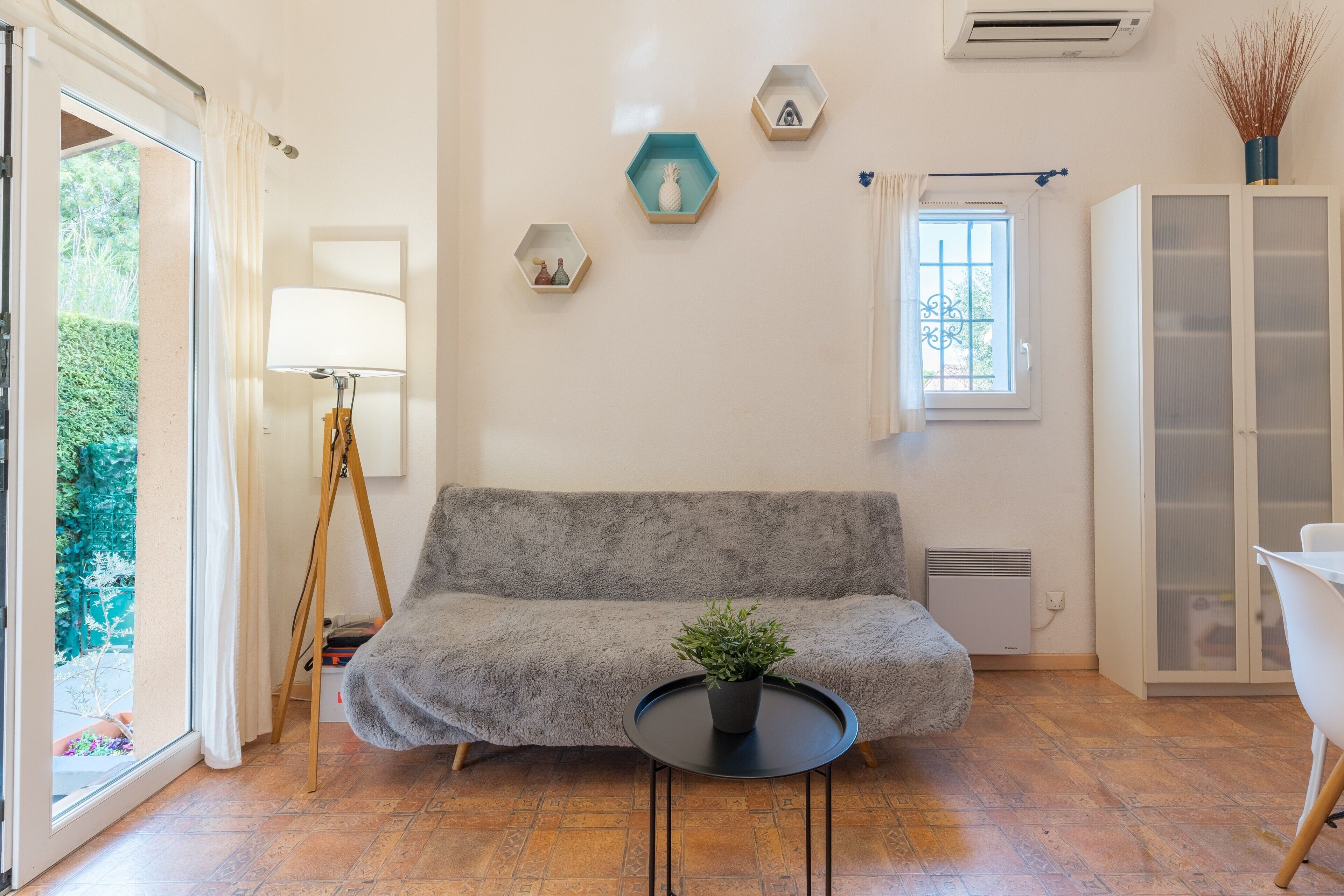 Ein gemütliches Zimmer mit grauem Sofa, einem kleinen runden Tisch mit Pflanze, einer Lampe und einem Fenster mit Vorhängen.